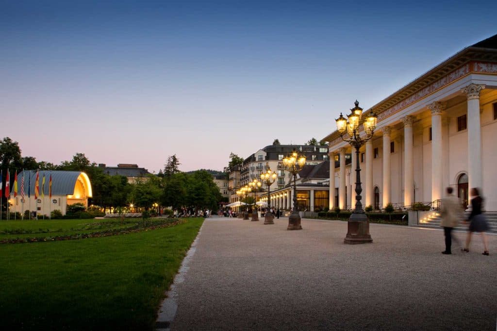 Een breed pad met sierlijke straatlantaarns leidt langs een groot neoklassiek gebouw met zuilen. Mensen wandelen er in de schemering langs en genieten van de dromerige Duitse sfeer. Kleurrijke vlaggen en een klein podium versieren de groene gazons en bomen in de buurt.