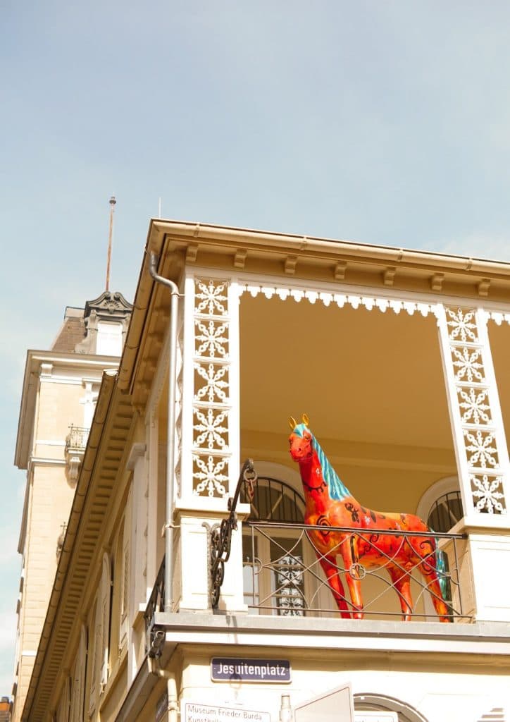 Een felrood geschilderd paardenbeeld met blauwe en gele accenten staat op het balkon van een crèmekleurig gebouw met sierlijk wit rasterwerk in dromerig Duitsland. Beneden staat op een straatnaambordje "Jesuitenplatz" tegen een helderblauwe lucht.