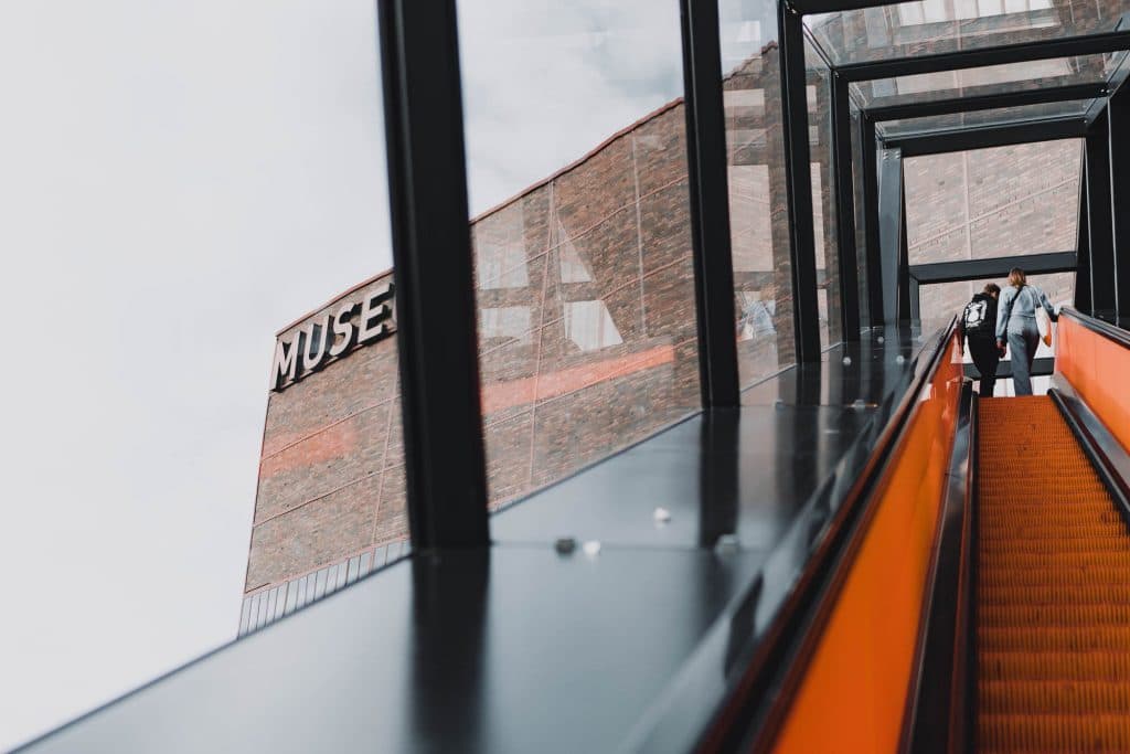 Twee mensen rijden op een oranje roltrap binnen een glazen omheining in Duisburg. Een bakstenen gebouw met het deels zichtbare woord "MUSE" staat op de achtergrond - een opvallende fotolocatie vanuit een lage hoek kijkend naar de lucht.