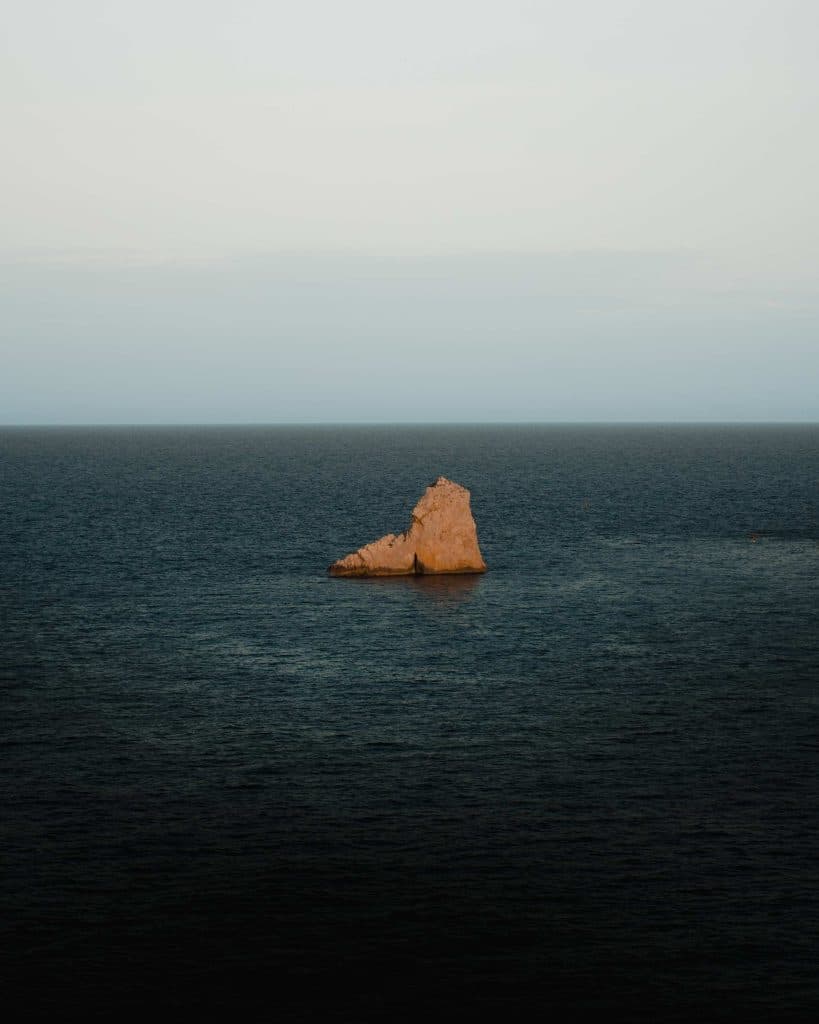 Een enkele grillige rotsformatie rijst op uit de donkerblauwe oceaan voor de Costa Brava onder een bleke, bewolkte hemel. Zonlicht verlicht de rots, contrasteert met de uitgestrekte, kalme zee en roept een gevoel van rustige afzondering op in deze hoek van Spanje.