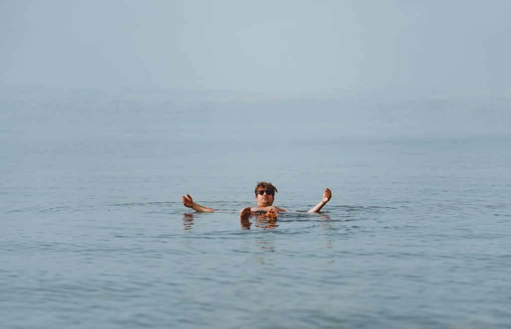 Een persoon met een zonnebril drijft kalm op zijn rug in een groot, rustig waterlichaam - een van de hoogtepunten van Jordanië - met armen en benen gedeeltelijk opgeheven boven het oppervlak en verre heuvels zichtbaar op de achtergrond.