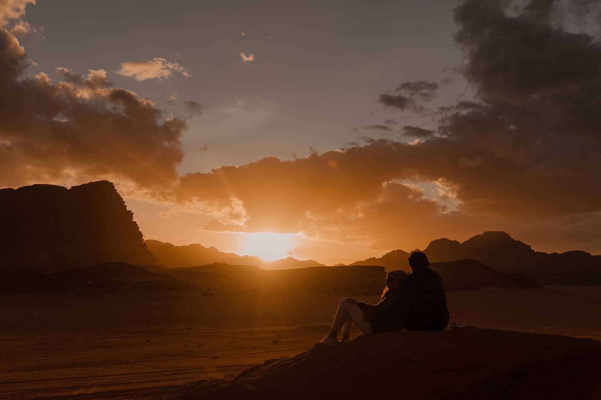 Twee mensen zitten dicht tegen elkaar aan op een rots, kijkend naar een dramatische zonsondergang over het woestijnlandschap van Jordanië, met silhouetten van bergen en wolken op de achtergrond-een magisch moment geïnspireerd door Jordaanse nomaden.