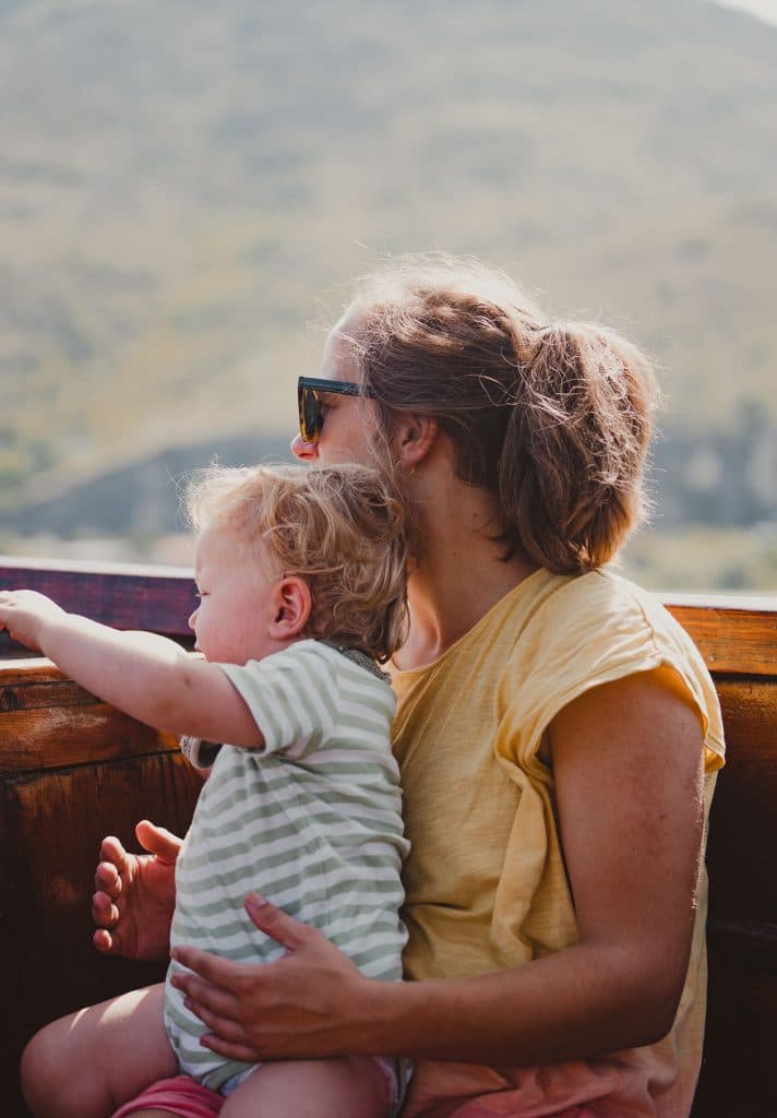 Een vrouw in een geel shirt en een zonnebril houdt een jong kind op haar schoot. Ze zitten buiten op een houten stoel, met zonovergoten groen en vage Balkanbergen op de achtergrond - een vredig moment tussen de mooiste landen.
