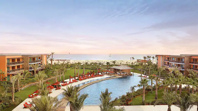 Een resort in Kaapverdie met palmbomen en rode loungestoelen omringt een groot, gebogen zwembad. Groene gazons leiden naar zandstranden en de oceaan, waar zeilboten in de verte dobberen onder een pastelkleurige hemel.