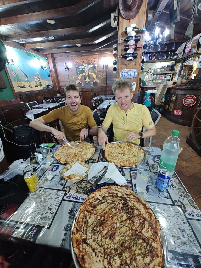 Twee lachende mannen in gele shirts zitten aan een restauranttafel met grote pizza's en drankjes, klaar om te eten. Het gezellige interieur heeft een houten inrichting, muurkunst en vintage memorabilia - perfect om reistips te delen of een reisblog te schrijven.