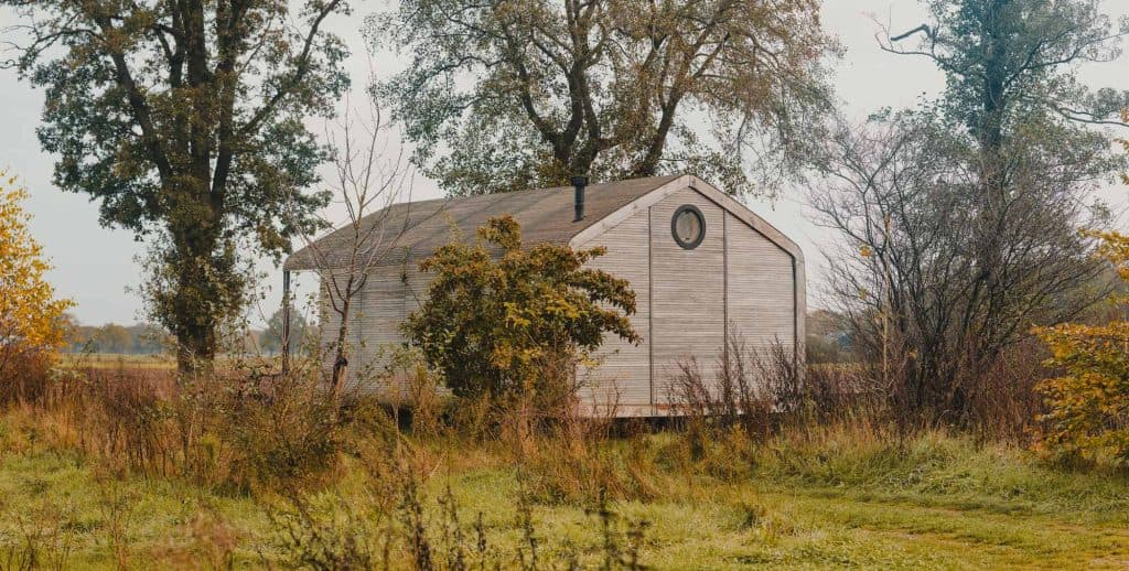 Een kleine, lichte houten hut met een rond raam staat omringd door hoog gras, struiken en bomen in een vredig, landelijk landschap - een van de echte hoogtepunten voor natuurliefhebbers die op zoek zijn naar somsookheimwee.