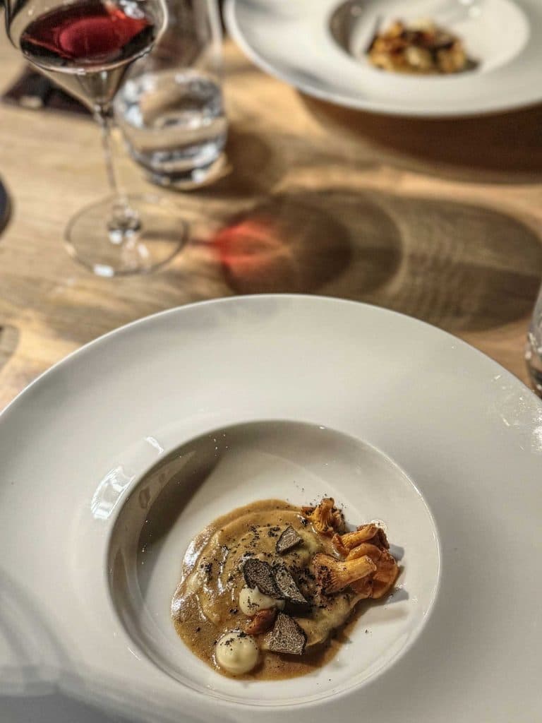 Een wit bord met een gastronomisch gerecht met ravioli overgoten met saus, champignons en geschaafde truffel. Perfect voor een weekendje Vlieland, een glas rode wijn en een ander gerecht op de zacht verlichte houten tafel op de achtergrond.