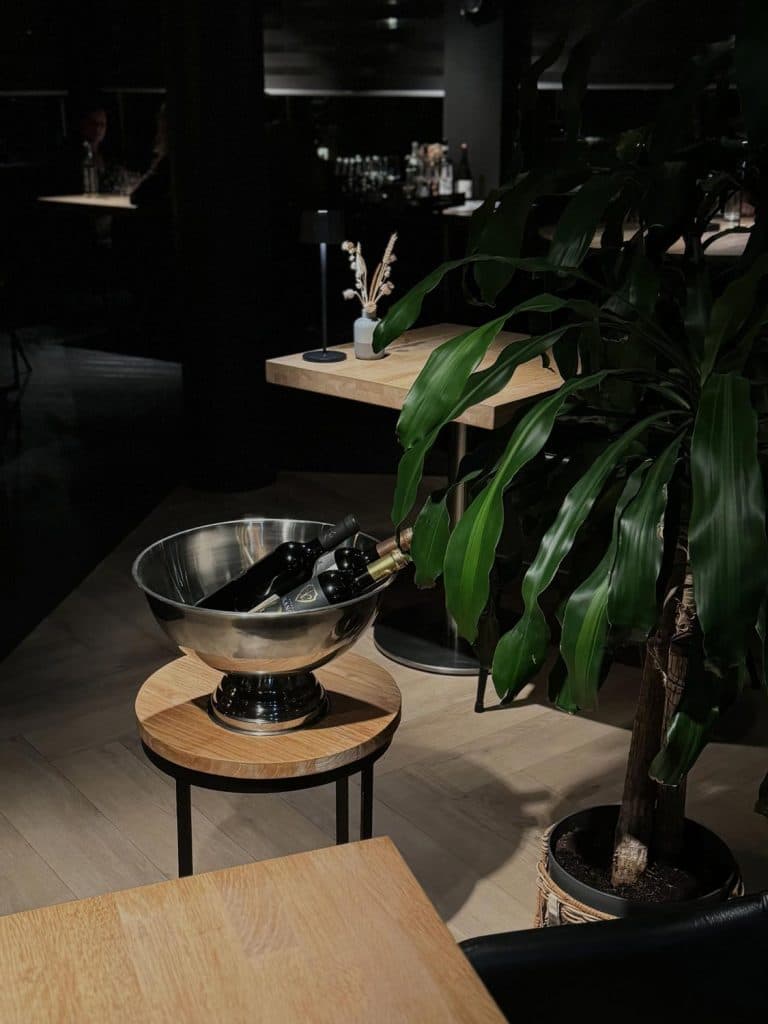Een schemerig verlicht restaurant op Vlieland, met een metalen wijnkoeler met twee flessen wijn op een kleine ronde tafel, omringd door houten tafels en een grote groene potplant - perfect voor een ontspannen Vlieland-weekendgevoel.