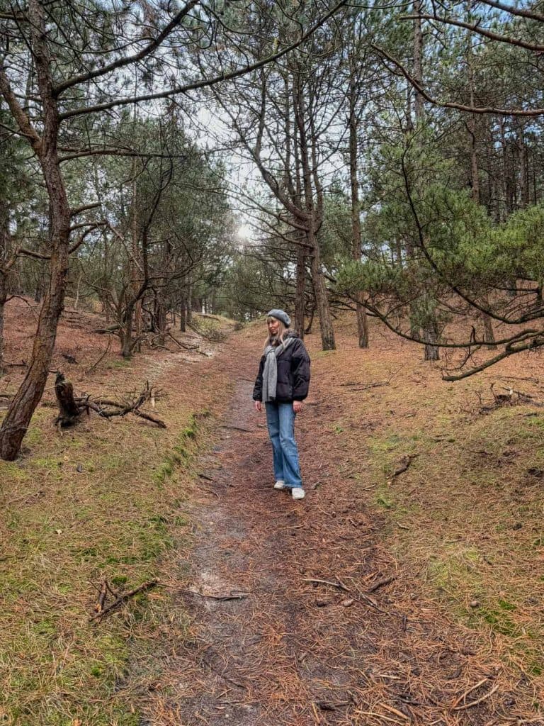 Een persoon met een jas, sjaal en muts staat op een met dennennaalden bedekt pad in een bos met hoge, slanke bomen en een bewolkte hemel boven hem. Dit geeft de rustige sfeer weer van een weekendje Vlieland.