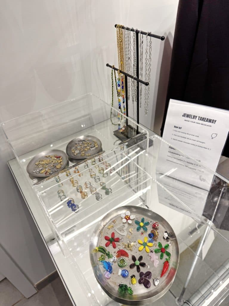 Een vitrinekast met kleurrijke juwelen op ronde dienbladen, kettingen die aan een rek hangen en een bordje "Jewelry Takeaway" op een glazen plank - perfect voor wie houdt van vintage shoppen in Hasselt.