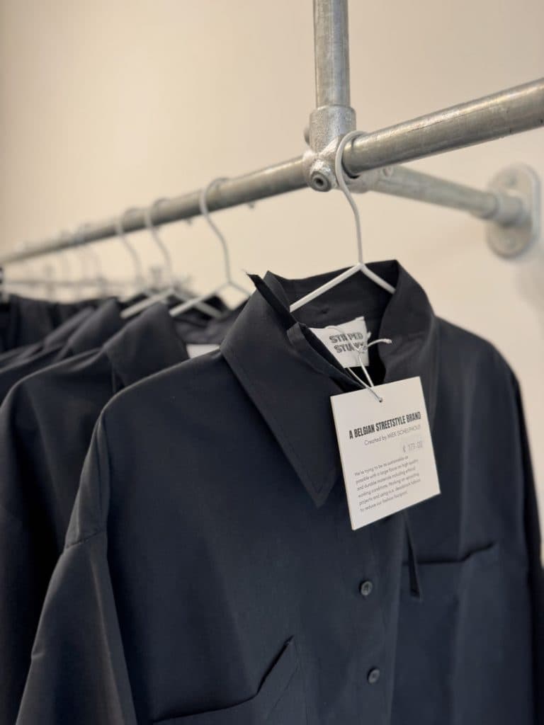 Een rij zwarte overhemden met knoopsluiting hangt aan een metalen kledingrek in Hasselt. Op een label aan de voorkant staat "STUDIO STADT" en informatie over het merk en de prijs - perfect voor vintage shoppen in Hasselt.