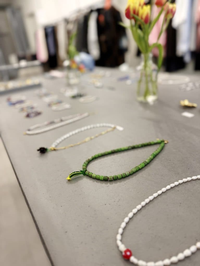 Kettingen en kralenjuwelen uitgestald op een grijze tafel, met vage vazen met tulpen en rekken met kleren op de achtergrond - perfect voor wie houdt van vintage shoppen in Hasselt.