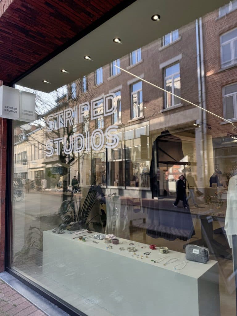 Een straatbeeld van een etalage met sieraden en modeartikelen in Hasselt. Op het uithangbord van de winkel staat "STRIPED STUDIOS" in grote witte letters, perfect voor wie houdt van vintage shoppen in Hasselt, met weerspiegelingen van de stad zichtbaar in het glas.