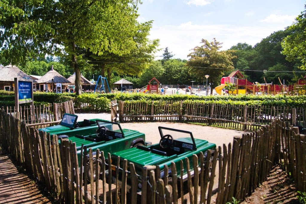 Een speeltuin met houten omheining, groene speelgoedauto's voor kinderen, schommels en kleurrijke klimstructuren - perfect voor een vakantie met kinderen tussen de bomen en het groen op een zonnige dag.