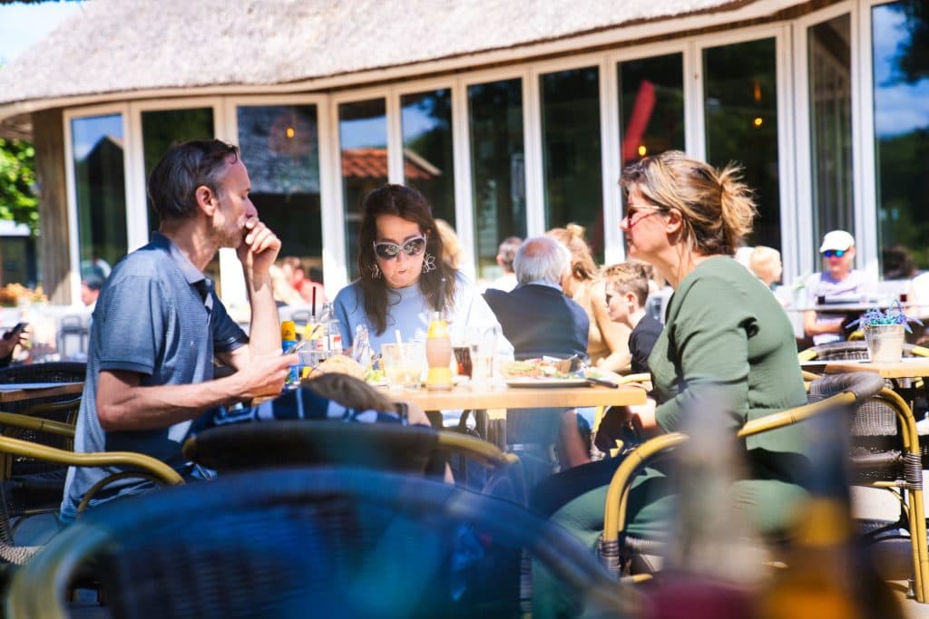 Drie mensen zitten aan een cafétafel buiten te eten en te praten over hun vakantie met kinderen in de Efteling. Het zonlicht schijnt op hen en op de achtergrond zijn andere eters zichtbaar. De sfeer is ontspannen en ongedwongen.