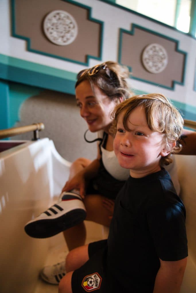 Een lachende jongen met krullend haar zit naast een vrouw, waarschijnlijk zijn moeder, tijdens een ritje in een pretpark - misschien geniet hij van een vakantie met kinderen na het krijgen van kaartjes voor de Efteling. De vrouw draagt een zonnebril op haar hoofd; beiden zien er blij en opgewonden uit.