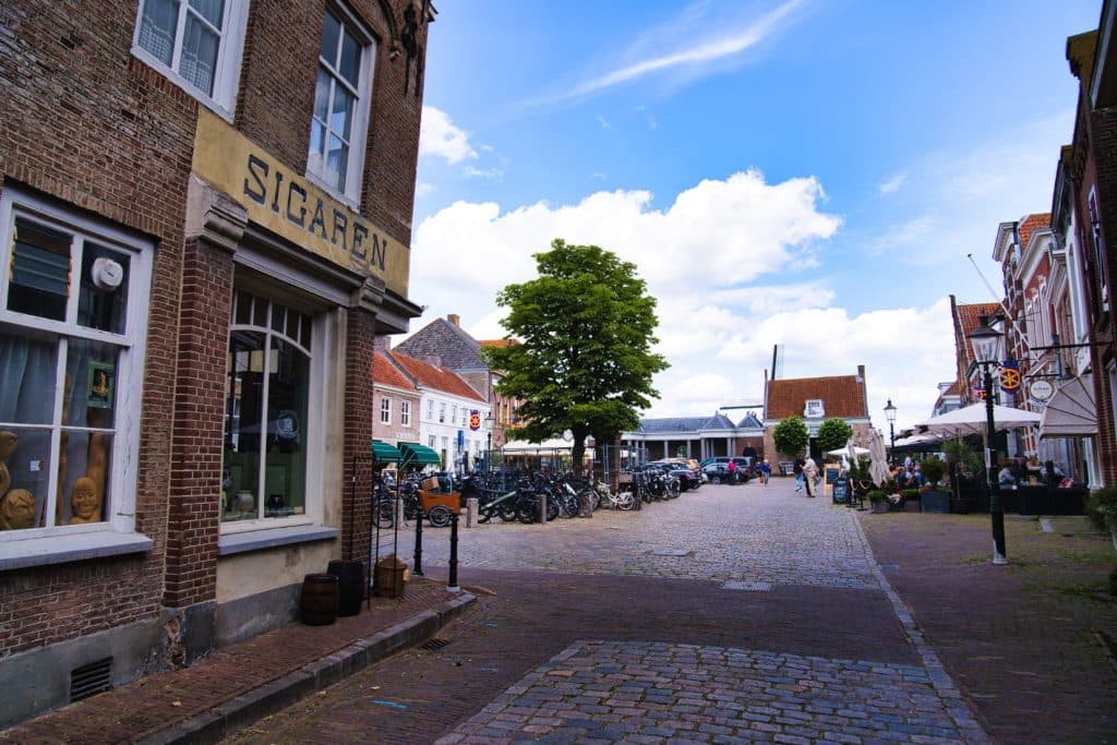 Een geplaveide straat in het schilderachtige Europese stadje Heusden met historische bakstenen gebouwen, een boom in het midden, fietsen geparkeerd langs de straat en mensen die flaneren bij terrasjes onder een gedeeltelijk bewolkte hemel.