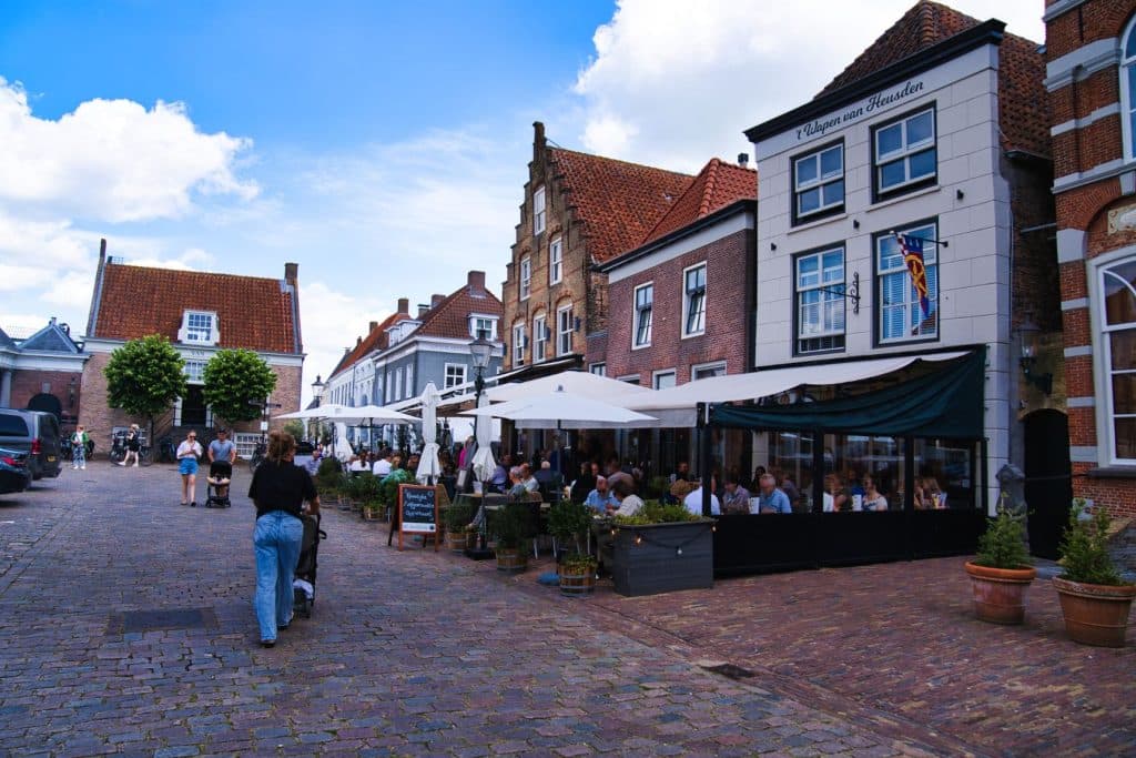 Mensen zitten aan buitentafels onder parasols in een café aan een geplaveide straat in historisch Heusden, terwijl anderen vlakbij wandelen of fietsen op een zonnige dag - perfect voor wie Heusden wil bezoeken na een leuk uitstapje naar de Efteling of Loonsche Land.