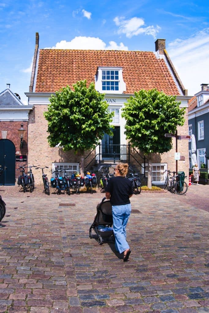 Een persoon duwt een kinderwagen over een geplaveide straat in de richting van een bakstenen gebouw met een pannendak, geflankeerd door lommerrijke bomen en geparkeerde fietsen - een typisch tafereel voor vakantie met kinderen in Nederland onder een blauwe lucht met verspreide wolken.
