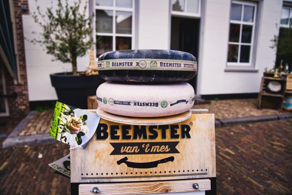 Twee grote wielen Beemster kaas liggen op een houten krat met de tekst "Beemster van 't mes" buiten een wit gebouw, met een ansichtkaart met eten - perfect om te spotten als je heusden bezoekt tijdens een vakantie met kinderen.