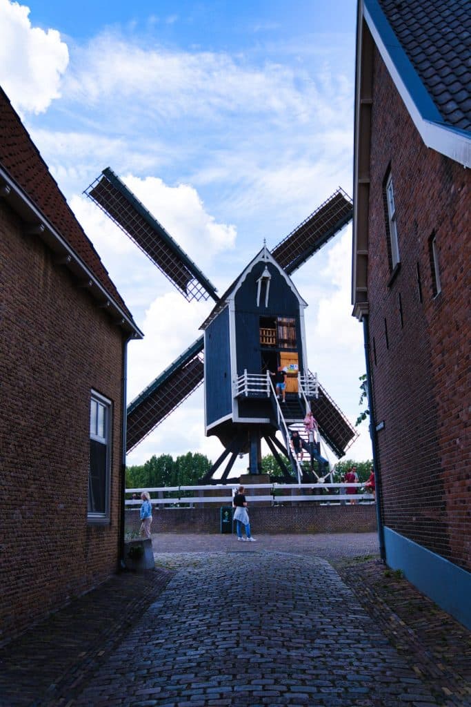 Een traditionele Nederlandse windmolen met donkere houten panelen staat tussen twee bakstenen gebouwen in Heusden, gezien vanuit een geplaveide steeg. Mensen lopen vlakbij onder een gedeeltelijk bewolkte blauwe lucht - perfect voor wie Heusden bezoekt of inspiratie zoekt voor wat doen in Nederland.