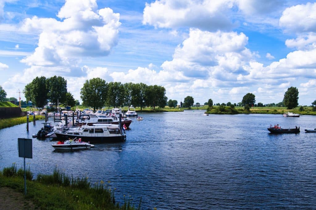 Een rivier met boten aangemeerd aan de met gras begroeide oever, omringd door groene bomen onder een blauwe lucht vol pluizige witte wolken. Perfect voor een vakantie met kinderen of voor wie zich afvraagt wat doen in Nederland-families samen genieten op het water.