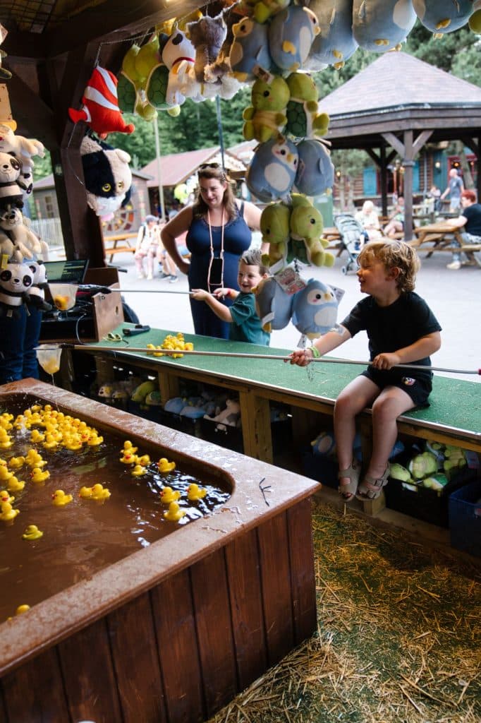 Twee kinderen spelen een eendenvisspel bij een kraampje op de buitenmarkt, omringd door hangende knuffeldierenprijzen, genietend van hun vakantie met kinderen. Een volwassen vrouw staat vlakbij hen te kijken terwijl mensen ontspannen aan picknicktafels op de achtergrond.