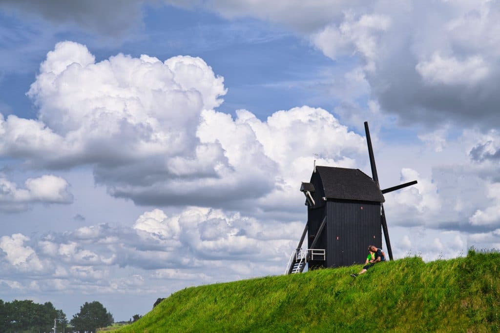 Een donkere houten windmolen staat op een met gras begroeide heuvel in Loonsche Land onder pluizige wolken. Een persoon in lichte kleding zit vlakbij en geniet van het schilderachtige uitzicht - een van de vele vredige momenten die je kunt beleven als je denkt aan wat doe je in Nederland.