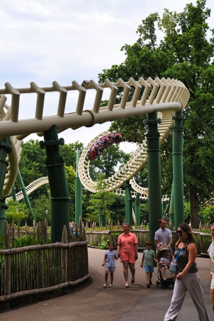 Gezinnen lopen over een pad onder een looping van een witte achtbaan die boven groene steunbalken draait in Loonsche Land, een park omringd door bomen en houten hekken - perfect voor wie zich afvraagt wat doen in Nederland. Een achtbaantreintje is zichtbaar op de baan.