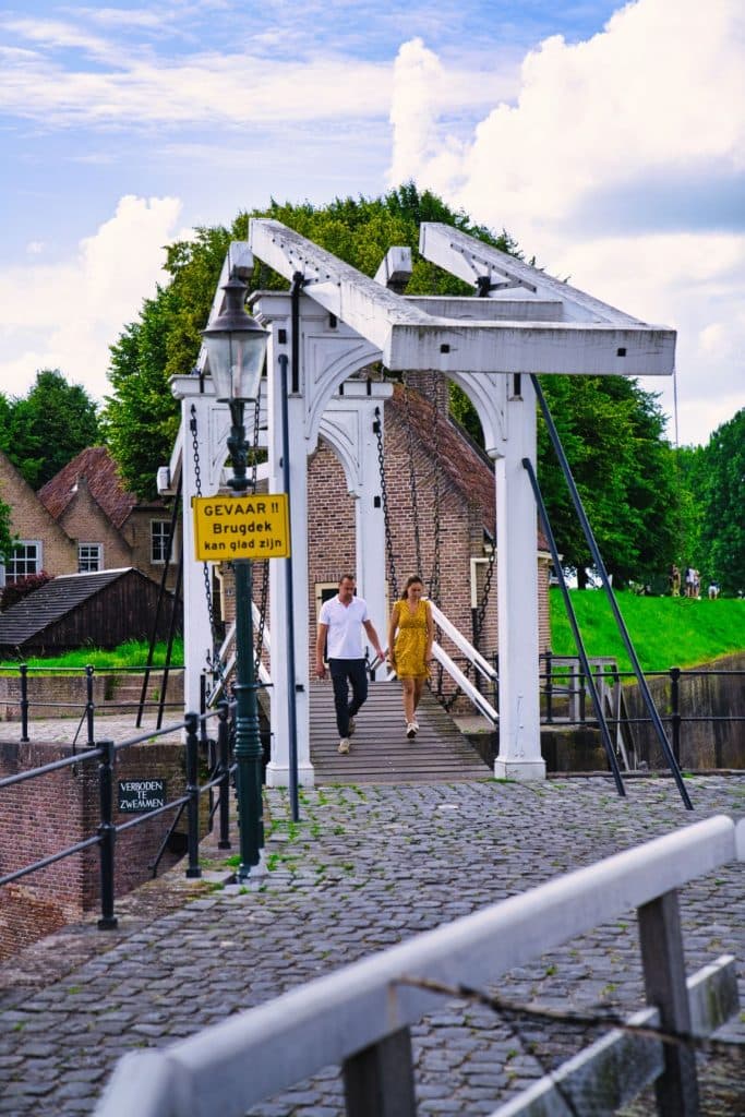 Een man en een vrouw lopen over een witte houten ophaalbrug over een keienpad in Heusden bezoeken. Op een geel bord op de brug staat: "GEVAAR!!! Brugdek kan blij zijn." Historische gebouwen en groen zijn zichtbaar op de achtergrond.