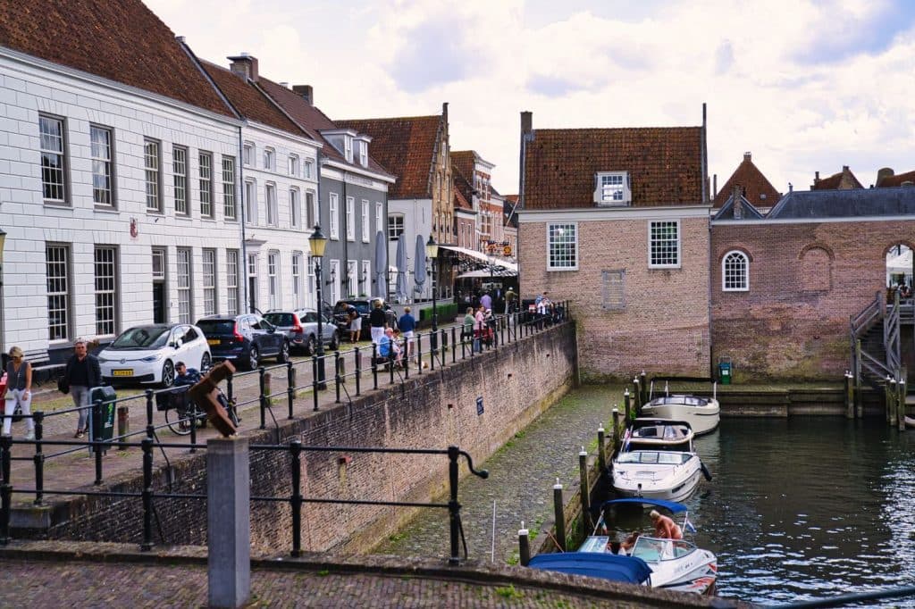 Een gracht met geparkeerde boten loopt langs een geplaveide straat met historische Europese gebouwen. Mensen flaneren over straat in dit charmante stadje - perfect voor wie op zoek is naar heusden bezoeken of ontdekken wat doen in Nederland.