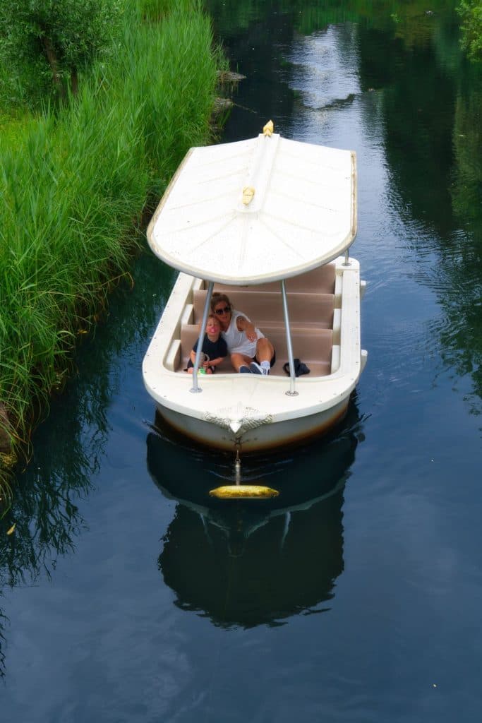 Twee mensen zitten samen op een klein wit bootje met een overkapping, drijvend op een rustige waterweg in de Efteling, omringd door groen gras en planten. Het water weerspiegelt de boot en de lucht erboven - perfect voor een vakantie met kinderen.
