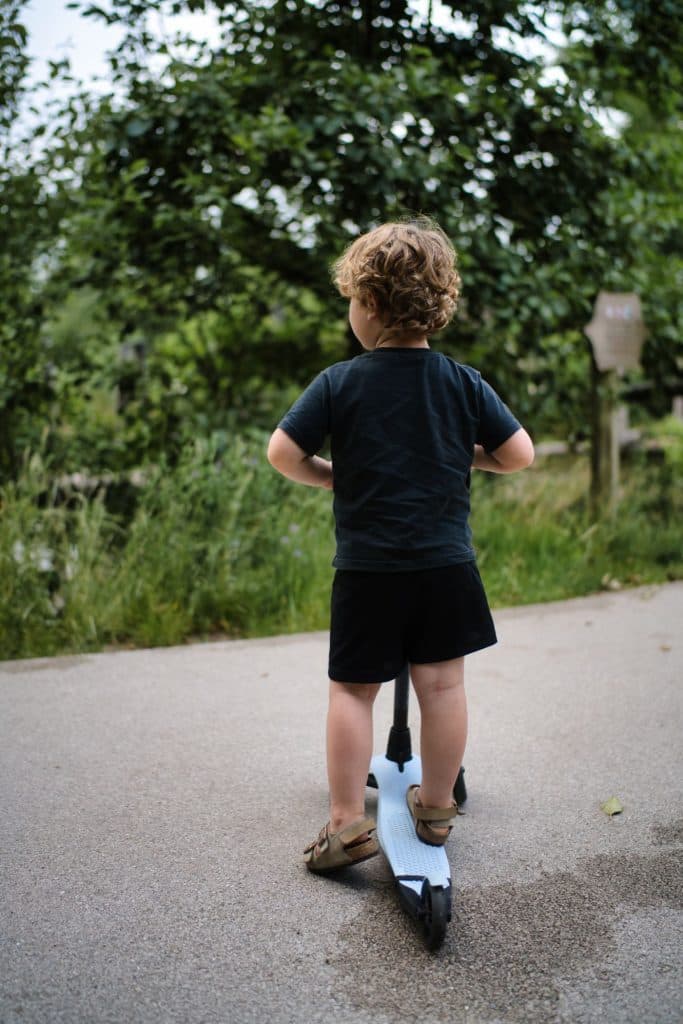 Een jong kind met krullend haar, gekleed in een zwart t-shirt, een zwarte korte broek en sandalen, staat op een step op een verhard pad omgeven door groen - perfect voor een vakantie met kinderen of nadat je kaartjes voor de Efteling hebt gebruikt. Het kind kijkt weg van de camera.