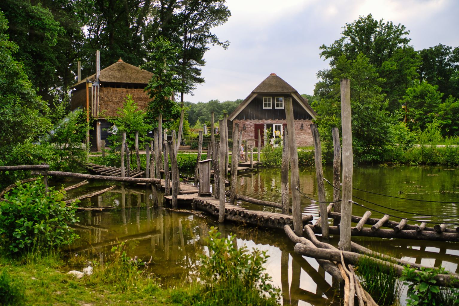 Een rustieke houten loopbrug steekt een vijver over en leidt naar een schilderachtig bakstenen huis met een rieten dak, omringd door weelderig groene bomen - perfect voor een vakantie met kinderen na een magische dag in de Efteling of het verkennen van wat te doen in Nederland.