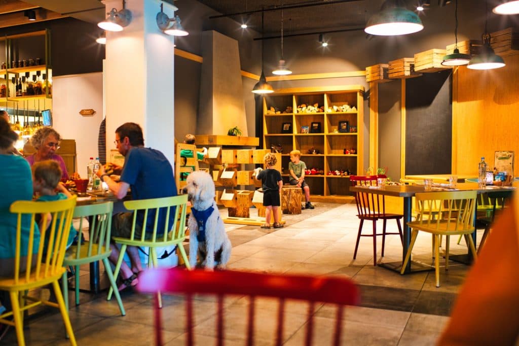 Een licht, modern restaurant met gezinnen die aan kleurrijke tafels dineren - perfect voor een maaltijd na het genieten van wat doen in Nederland of een bezoek aan de Efteling. Kinderen spelen in de buurt terwijl warme verlichting en een witte hond bijdragen aan de gezellige, levendige sfeer.