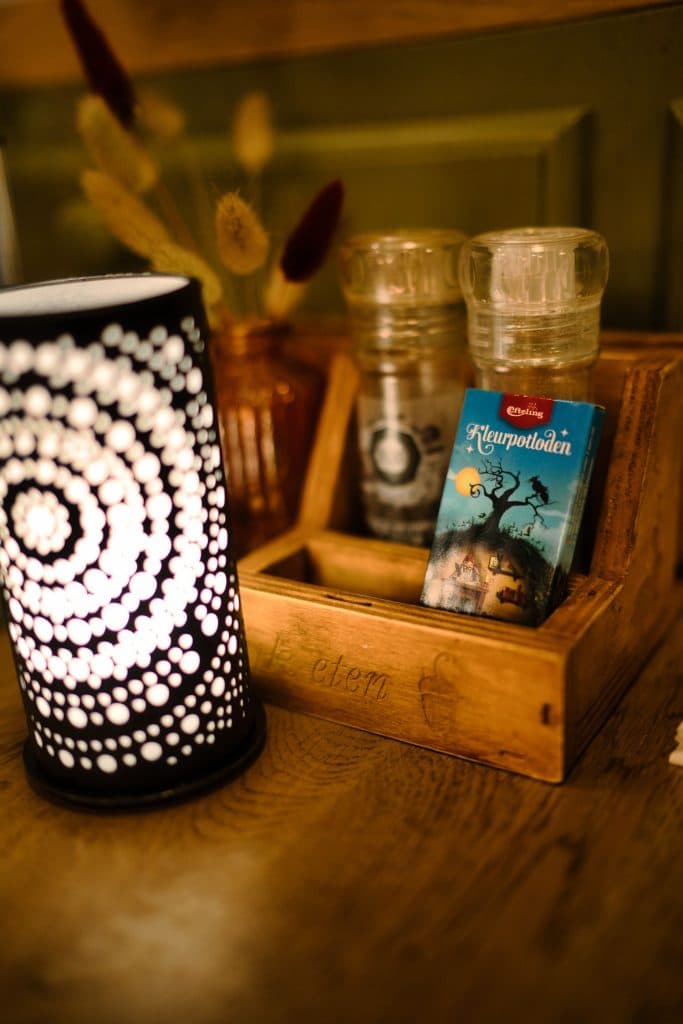 Een houten dienblad bevat een glazen peper- en zoutstel en een blauwe kaart met een boom en een maan. Naast het dienblad staat een zwart-witte lamp met cirkelvormige patronen, gedroogde bloemen in een klein vaasje, perfect na een bezoek aan de Efteling of Loonsche Land.