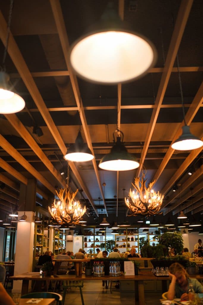 Een warm verlicht restaurantinterieur met houten plafondbalken, geweivormige kroonluchters en hanglampen - perfect voor een vakantie met kinderen na een dag verkennen van wat te doen in Nederland. Mensen dineren terwijl planken met planten en decor langs de muren hangen.