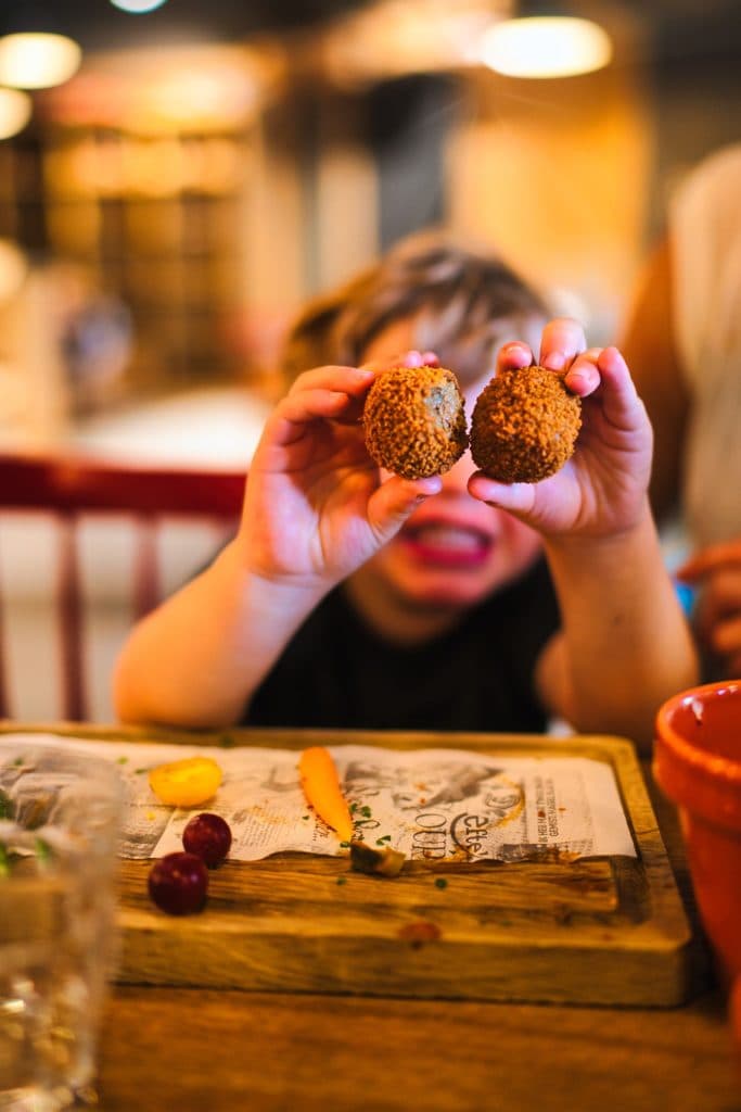 Een jong kind houdt twee ronde, gepaneerde snacks omhoog naar de camera terwijl het aan een tafel zit tijdens een vakantie met kinderen, met eten en een houten dienblad voor zich. De achtergrond is onscherp, waardoor het kind en de snacks worden uitgelicht.