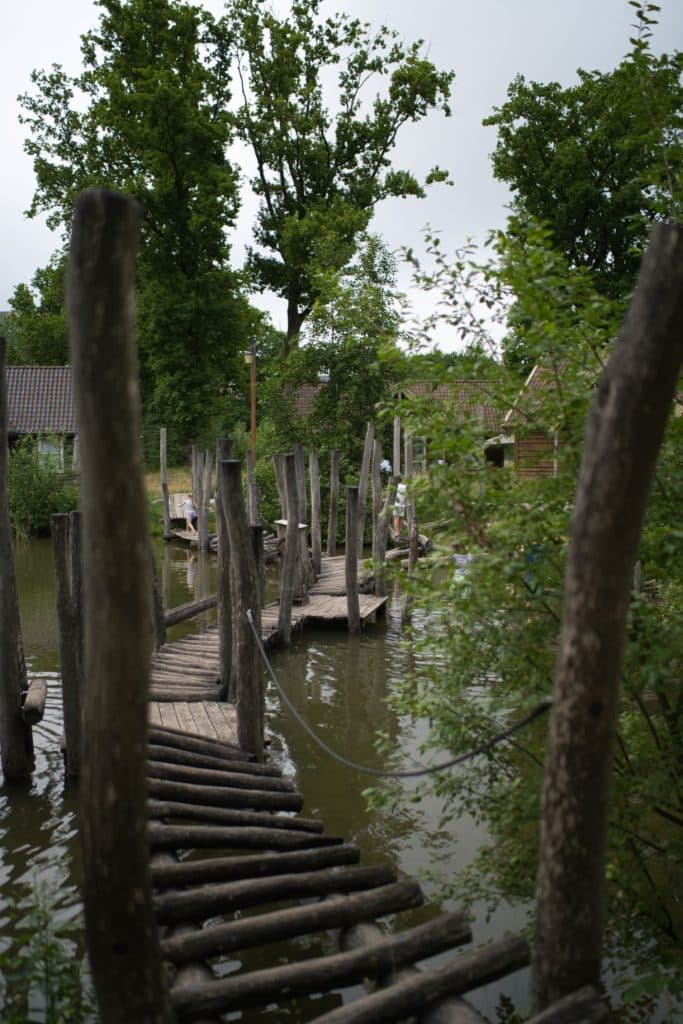 Een rustieke houten loopbrug met ongelijke planken en hoge palen strekt zich uit over een vijver, omringd door weelderige groene bomen - een serene plek die perfect is voor een vakantie met kinderen op een bewolkte dag.
