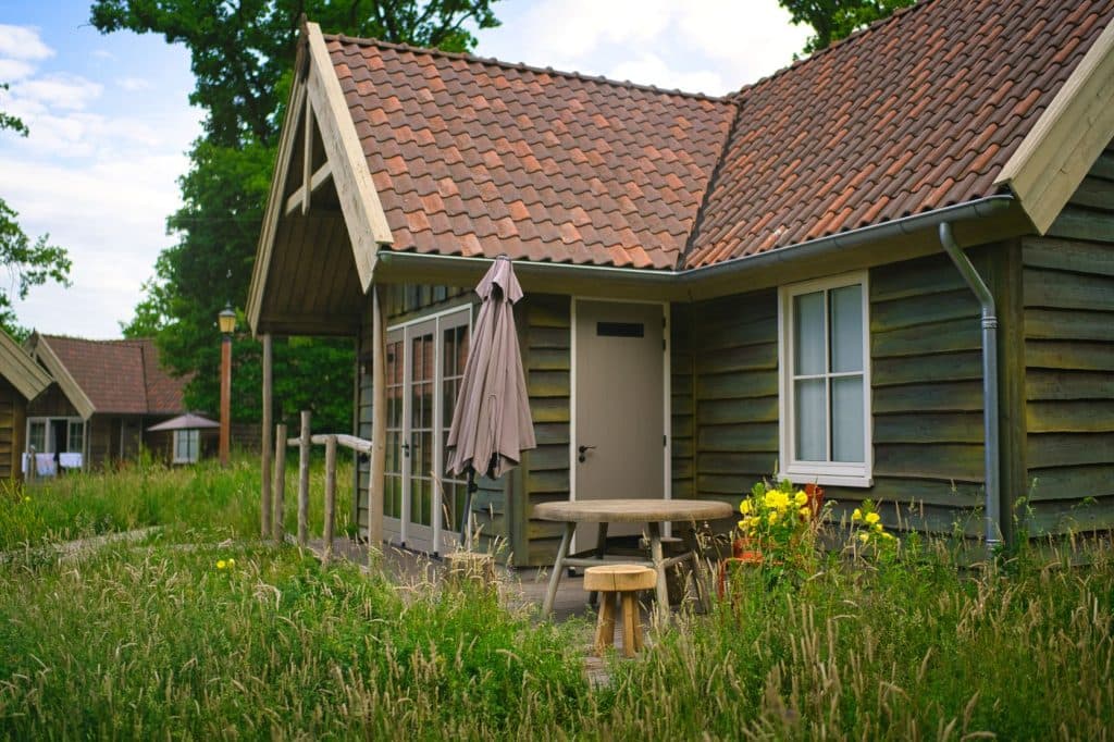 Een kleine houten hut met een rood pannendak staat in een grasveld, met een ronde houten tafel, krukjes en een gesloten parasol buiten. Weelderige groene bomen omringen de vredige omgeving - perfect om te ontspannen nadat je heusden hebt bezocht.