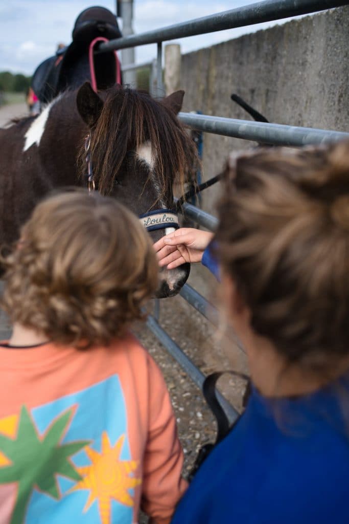 Een kind en een volwassene aaien een zwart-witte pony genaamd Loulou door een metalen hek in Loonsche Land. Het kind draagt een oranje shirt met kleurrijke sterrendessins, terwijl de volwassene met opgebonden bruin haar naast hen glimlacht.