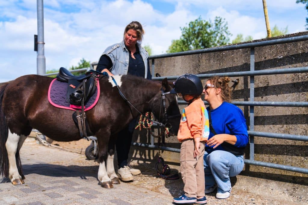 Een jong kind met een helm op staat naast een kleine zwart-witte pony, zachtjes vastgehouden door een lachende volwassene die vlakbij knielt. Beleef dit vrolijke moment tijdens je vakantie met kinderen bij Loonsche Land in een zonnige buitenomgeving met een metalen hek.