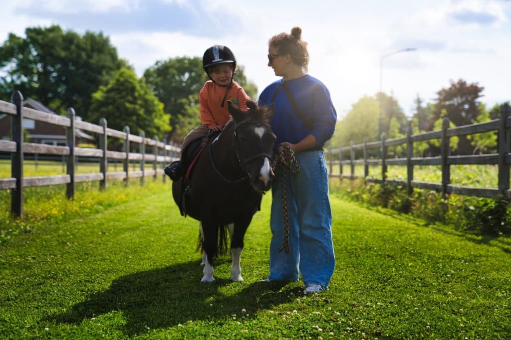 Een jong kind met een helm op berijdt een kleine zwarte pony, geleid door een volwassen vrouw die de teugels vasthoudt op een zonnig, grasachtig pad omzoomd door houten hekken en bomen - perfect voor een vakantie met kinderen in Nederland.