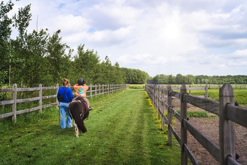 Een kind met een helm op rijdt op een klein paard over een graspad in het Loonsche Land van de Efteling, omzoomd met houten hekken, vergezeld door een volwassene die naast hen loopt onder een halfbewolkte hemel.