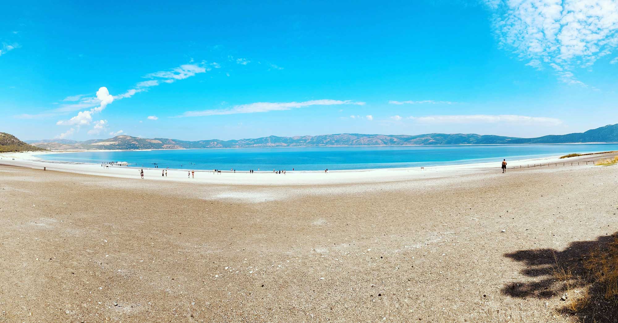 Een breed zandstrand leidt naar een turquoise meer onder een helderblauwe hemel met verspreide wolken. Mensen wandelen langs de oever en bergen rijzen in de verte op aan de horizon - perfect voor vakantie in Turkije en natuurliefhebbers.