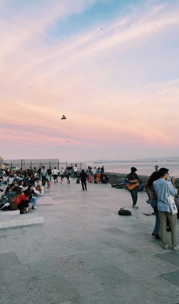Mensen verzamelen zich op een promenade aan zee in Lissabon bij zonsondergang, sommigen zitten op bankjes en anderen staan. Een straatmuzikant speelt gitaar terwijl vogels in de pastelkleurige lucht vliegen - een levendig, ontspannen tafereel tussen de must-dos van de stad.