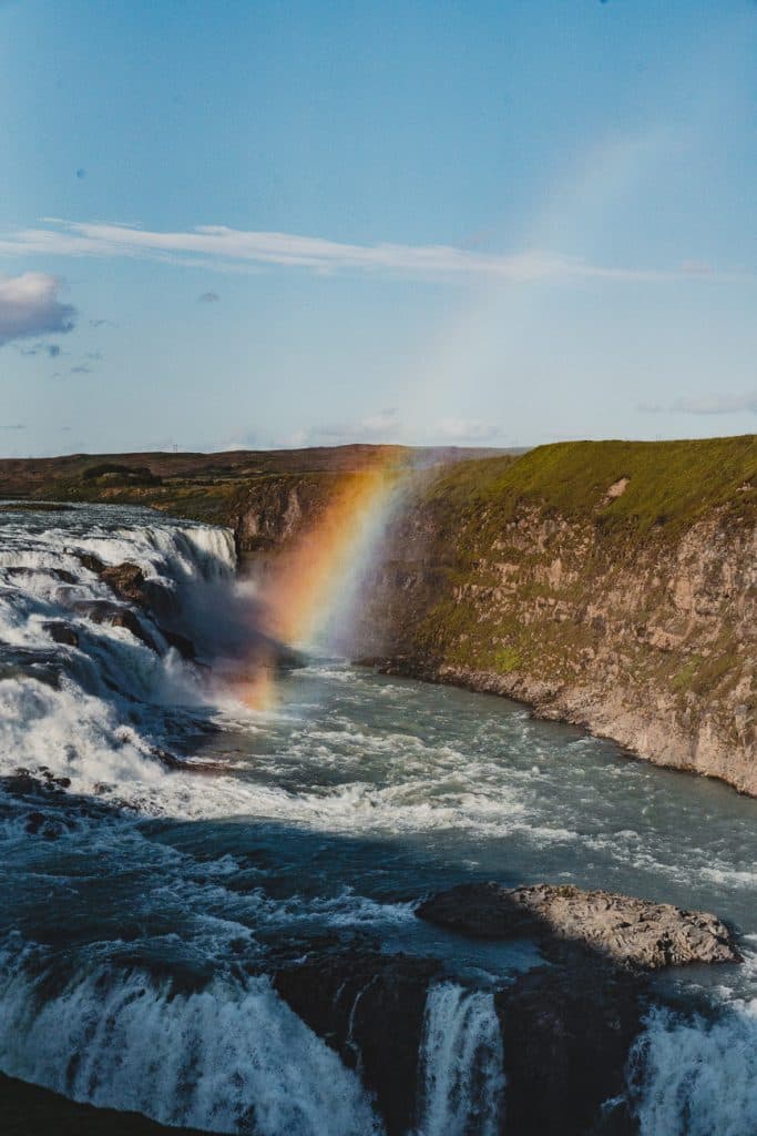Een levendige regenboog verschijnt boven een krachtige waterval in IJsland, met water dat in een rotsachtige rivier stroomt. Weelderige groene kliffen, symbolen van groei, omzomen het stromende water onder een helderblauwe hemel.