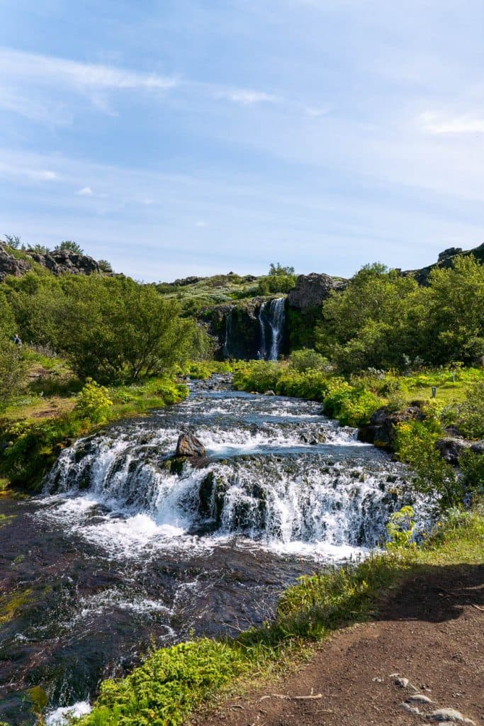 Kleine watervallen stromen door een weelderige groene vegetatie onder een blauwe hemel, met een grotere waterval op de achtergrond en een zandpad dat naar dit serene IJsland-landschap leidt.
