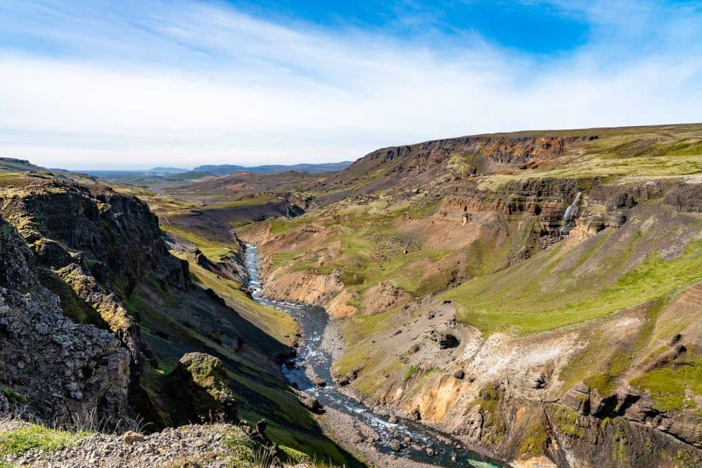 Een weids uitzicht op een rotsachtige canyon in IJsland waar een kronkelende rivier doorheen stroomt, omringd door groene groeiplekken en ruige heuvels onder een blauwe lucht met lichte wolken.