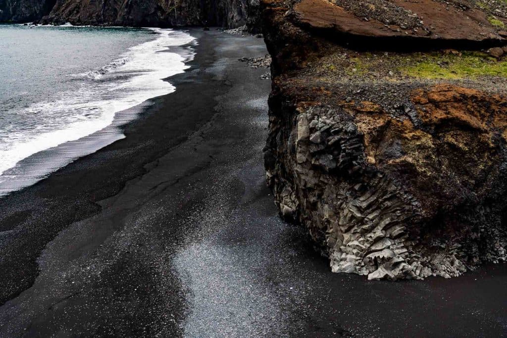 Een ruige klif rijst op boven een zwart zandstrand in IJsland, met donker vulkaanzand dat zich uitstrekt langs de kust en golven die zachtjes aanspoelen, en de rauwe schoonheid en gestage groei van de natuur laten zien.